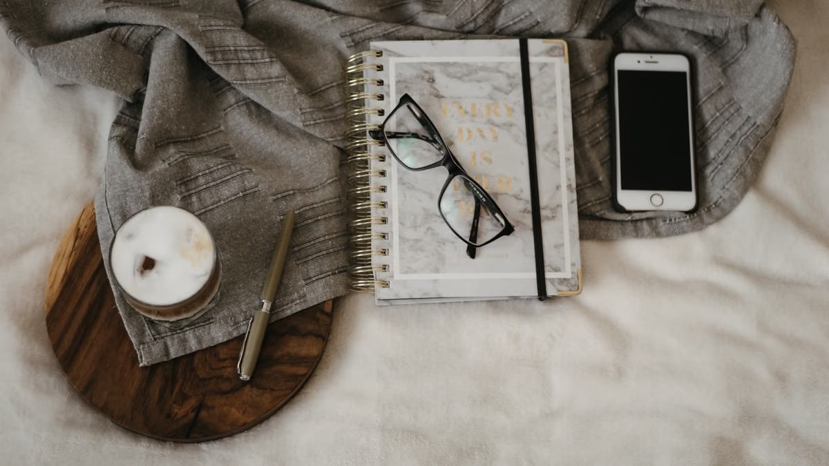A cozy flat lay featuring a notebook, glasses, coffee, smartphone, and pen on a soft fabric.